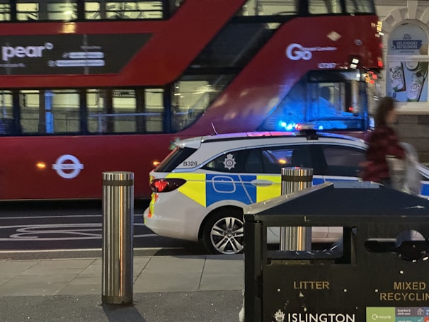 A red double-decker bus is moving past a parked police car with blue and yellow markings. A recycling bin is visible in the foreground, and a person is walking near the edge of the frame. The scene takes place in an urban setting during the evening, with dim lighting and artificial lights cast on the street.
