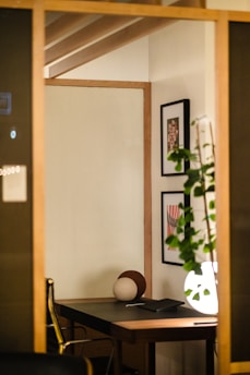 A cozy, modern interior space featuring a wooden desk with a decorative sphere placed on it. The area is warmly lit, creating an inviting atmosphere. On the wall, framed artwork adds a touch of sophistication, and a potted plant contributes a natural element.