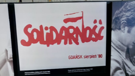 The image features a poster with the word 'Solidarność' in bold red letters, symbolizing a flag, against a white background. Below the word, it reads 'GDAŃSK sierpień '80'. To the right, there is a grayscale photograph of a person.