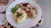 A plate with Swedish meatballs covered in creamy gravy, surrounded by mashed potatoes with herbs on top, green peas, pickled cucumbers, and lingonberry sauce. A dessert roll is partially visible to the side, with a napkin that reads 'IKEA Museum' placed nearby.