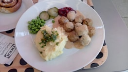 A plate with Swedish meatballs covered in creamy gravy, surrounded by mashed potatoes with herbs on top, green peas, pickled cucumbers, and lingonberry sauce. A dessert roll is partially visible to the side, with a napkin that reads 'IKEA Museum' placed nearby.