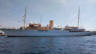 A large, classic yacht with two masts and a light brown and white color scheme is seen resting on calm, blue water under a clear sky. The yacht has traditional design elements, including a tall funnel and multiple decks.