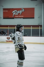 A hockey player wearing a white and black uniform with the number 23 stands on an ice rink. The player is equipped with a helmet, gloves, and skates. The background includes boards and advertisements with a large red sign overhead. Another player is visible in the background, also in hockey gear.