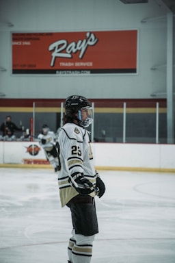A hockey player wearing a white and black uniform with the number 23 stands on an ice rink. The player is equipped with a helmet, gloves, and skates. The background includes boards and advertisements with a large red sign overhead. Another player is visible in the background, also in hockey gear.