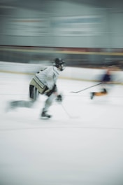 A dynamic and blurred image captures two ice hockey players in motion on the rink, one wearing a white and black uniform. The background features the indistinct outlines of a hockey arena with bright lights.