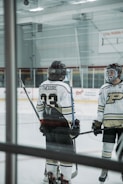 Two hockey players standing on an ice rink, wearing helmets and uniforms with numbers and letters visible on their backs. They both hold hockey sticks and are engaged in conversation or preparation. The rink features clear barriers and advertisements on the walls.