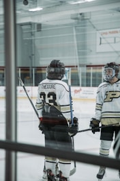 Two hockey players standing on an ice rink, wearing helmets and uniforms with numbers and letters visible on their backs. They both hold hockey sticks and are engaged in conversation or preparation. The rink features clear barriers and advertisements on the walls.