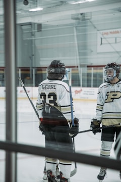 Two hockey players standing on an ice rink, wearing helmets and uniforms with numbers and letters visible on their backs. They both hold hockey sticks and are engaged in conversation or preparation. The rink features clear barriers and advertisements on the walls.