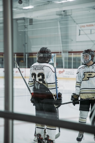 Two hockey players standing on an ice rink, wearing helmets and uniforms with numbers and letters visible on their backs. They both hold hockey sticks and are engaged in conversation or preparation. The rink features clear barriers and advertisements on the walls.