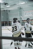 Several ice hockey players are gathered on the rink, wearing white and black uniforms. One player's jersey prominently displays the name 'Theodore' and the number 23. The players are equipped with helmets and hockey sticks, with a partially visible rink in the background.