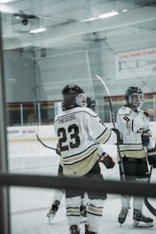 Several ice hockey players are gathered on the rink, wearing white and black uniforms. One player's jersey prominently displays the name 'Theodore' and the number 23. The players are equipped with helmets and hockey sticks, with a partially visible rink in the background.