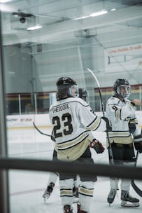 Several ice hockey players are gathered on the rink, wearing white and black uniforms. One player's jersey prominently displays the name 'Theodore' and the number 23. The players are equipped with helmets and hockey sticks, with a partially visible rink in the background.