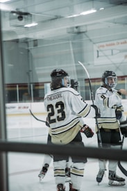 Several ice hockey players are gathered on the rink, wearing white and black uniforms. One player's jersey prominently displays the name 'Theodore' and the number 23. The players are equipped with helmets and hockey sticks, with a partially visible rink in the background.
