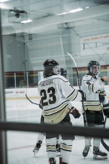 Several ice hockey players are gathered on the rink, wearing white and black uniforms. One player's jersey prominently displays the name 'Theodore' and the number 23. The players are equipped with helmets and hockey sticks, with a partially visible rink in the background.
