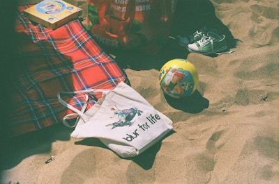 A plaid red and blue blanket spread on sandy ground is partially covered by a white tote bag with a print of a person, alongside a yellow ball featuring colorful cartoons. Nearby, there are sneakers and a red shopping bag, as well as a book with a bright cover.