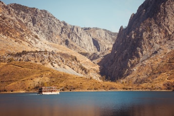 A scenic view of a large, calm lake set against a backdrop of rugged, rocky mountains. A tourist boat is sailing on the lake, leaving a gentle wake. The landscape is bathed in warm sunlight, highlighting the earthy tones of the cliffs and hills surrounding the water.