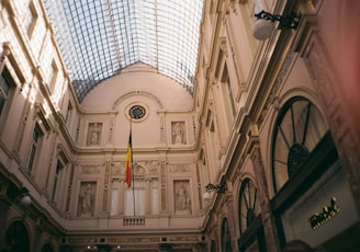 A grand architectural interior space with a high, ornate ceiling made of glass and iron. The walls are adorned with statues and decorative elements. A Belgian flag is prominently displayed in the center.