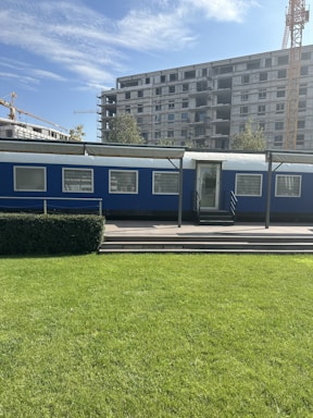 A blue portable cabin with multiple windows is set in front of a large construction site featuring a multi-story building. Cranes are visible in the background, and the area is surrounded by greenery, including a grassy field and trimmed bushes. The sky is clear with a few clouds.