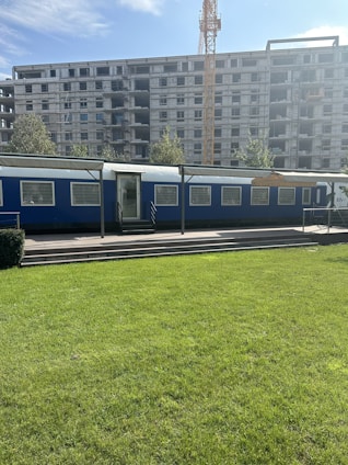 A construction site with a large unfinished building under development. In the forefront, there's a blue temporary office or trailer set against the backdrop of a green lawn. A crane towers in the background, indicating ongoing construction activities.