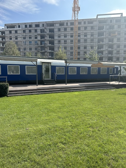 A construction site with a large unfinished building under development. In the forefront, there's a blue temporary office or trailer set against the backdrop of a green lawn. A crane towers in the background, indicating ongoing construction activities.