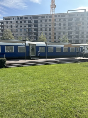 A construction site with a large unfinished building under development. In the forefront, there's a blue temporary office or trailer set against the backdrop of a green lawn. A crane towers in the background, indicating ongoing construction activities.