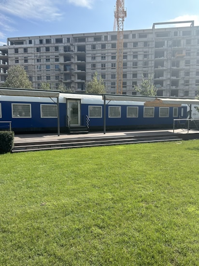 A construction site with a large unfinished building under development. In the forefront, there's a blue temporary office or trailer set against the backdrop of a green lawn. A crane towers in the background, indicating ongoing construction activities.