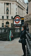 A person is standing near an entrance to the London Underground, specifically Bank Station. The person appears to be using a phone while leaning against a wall. The architecture in the background includes a large, classical building with tall columns.