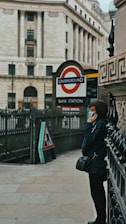 A person is standing near an entrance to the London Underground, specifically Bank Station. The person appears to be using a phone while leaning against a wall. The architecture in the background includes a large, classical building with tall columns.