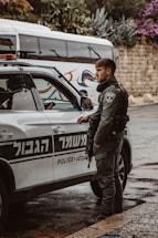 A police officer in tactical gear stands next to a marked police vehicle. The vehicle has signage in both Hebrew and English. In the background, there is a bus and stone wall partially covered by various plants and flowers.