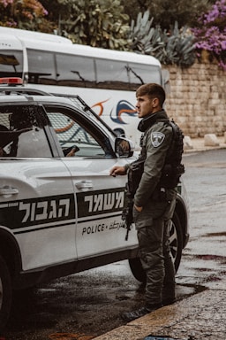 A police officer in tactical gear stands next to a marked police vehicle. The vehicle has signage in both Hebrew and English. In the background, there is a bus and stone wall partially covered by various plants and flowers.