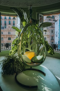 A modern, green-themed room featuring a hanging chair with a unique, nature-inspired design. The chair is surrounded by large, lush indoor plants and set against a backdrop of open windows showcasing an urban city view. The seat includes vibrant green and yellow cushions, enhancing the natural motif.