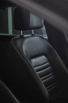 A close-up view of a car seat made of dark leather. The seat has detailed stitching and is equipped with a headrest supported by two metal rods. The sleek surface of the leather reflects light, emphasizing its texture and quality.