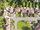 An aerial view of a residential area featuring houses with red-tiled roofs, paved driveways, and neatly maintained gardens. The arrangement of homes follows a curved street layout with parked cars visible in driveways and on the street. Surrounding the houses are lush green lawns and trees.