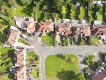 An aerial view of a residential area featuring houses with red-tiled roofs, paved driveways, and neatly maintained gardens. The arrangement of homes follows a curved street layout with parked cars visible in driveways and on the street. Surrounding the houses are lush green lawns and trees.