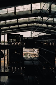 A modern architectural space with large glass walls and geometric structures, capturing a view of both interior and exterior environments. The interior features multiple floors with visible walkways and railings, while the exterior includes an industrial or urban setting in the distance.