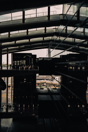 A modern architectural space with large glass walls and geometric structures, capturing a view of both interior and exterior environments. The interior features multiple floors with visible walkways and railings, while the exterior includes an industrial or urban setting in the distance.