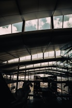 A modern architectural interior with large, expansive windows revealing a cloudy sky. The ceiling features a geometric pattern with support beams. Reflections of the exterior and other parts of the building can be seen on the glass surfaces.