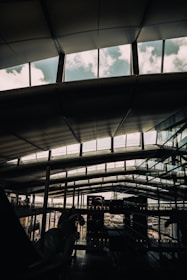 A modern architectural interior with large, expansive windows revealing a cloudy sky. The ceiling features a geometric pattern with support beams. Reflections of the exterior and other parts of the building can be seen on the glass surfaces.