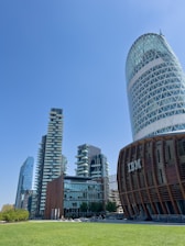 A modern urban landscape with several high-rise buildings featuring unique architectural designs. One building has a cylindrical shape with a lattice pattern, while others are tall with staggered balconies and glass exteriors. In the foreground, there is a well-maintained green lawn.