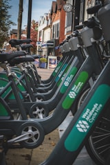 A row of bicycles lined up in a cycle hire station along a pedestrian street with a variety of shops and buildings in the background. The bicycles are part of the West Midlands Cycle Hire system and are parked on a paved walkway. The surrounding architecture features red brick and wood-framed structures, indicating a mix of modern and traditional design. A clock is visible on one of the buildings, and autumn trees can be seen nearby.