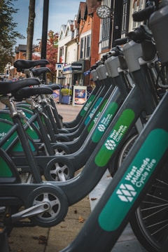 A row of bicycles lined up in a cycle hire station along a pedestrian street with a variety of shops and buildings in the background. The bicycles are part of the West Midlands Cycle Hire system and are parked on a paved walkway. The surrounding architecture features red brick and wood-framed structures, indicating a mix of modern and traditional design. A clock is visible on one of the buildings, and autumn trees can be seen nearby.