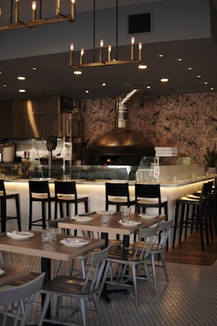 A cozy and modern restaurant interior with wooden tables and chairs arranged in a neat order. The bar area features high chairs with a well-lit counter. A wood-fired oven is prominently built into the background, giving the space a warm ambiance. Hanging light fixtures provide a soft glow above.