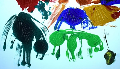 An abstract composition with textured paint splatters in various colors, including green, blue, red, and yellow. The paint appears thick and cracked, creating a dynamic and expressive visual effect against a white background.