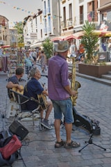 A lively street scene with musicians performing for passersby. The men are playing guitars and a saxophone, set on a cobblestone street with surrounding shops and colorful bunting overhead. People are walking and browsing nearby stalls, contributing to a vibrant atmosphere.