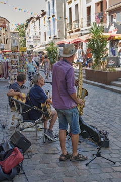 A lively street scene with musicians performing for passersby. The men are playing guitars and a saxophone, set on a cobblestone street with surrounding shops and colorful bunting overhead. People are walking and browsing nearby stalls, contributing to a vibrant atmosphere.