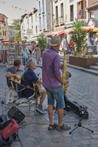 A lively street scene with musicians performing for passersby. The men are playing guitars and a saxophone, set on a cobblestone street with surrounding shops and colorful bunting overhead. People are walking and browsing nearby stalls, contributing to a vibrant atmosphere.