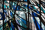 Intricate stained glass artwork displaying abstract geometric patterns with intersecting lines and vibrant hues. The glass is primarily composed of shades of blue, green, and white, with sections separated by dark leading, creating striking contrasts and an overall aesthetic of dynamic motion.