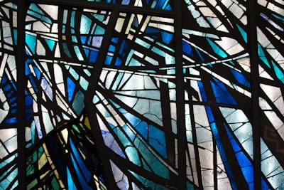 Intricate stained glass artwork displaying abstract geometric patterns with intersecting lines and vibrant hues. The glass is primarily composed of shades of blue, green, and white, with sections separated by dark leading, creating striking contrasts and an overall aesthetic of dynamic motion.