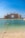A grand, luxurious hotel with distinctive architecture overlooks the calm blue waters of the sea. The pale sandy beach leads to clear, turquoise water with a line of buoys marking the swimming area. The sky is clear and bright, enhancing the overall elegance and serenity of the scene.