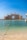 A grand, luxurious hotel with distinctive architecture overlooks the calm blue waters of the sea. The pale sandy beach leads to clear, turquoise water with a line of buoys marking the swimming area. The sky is clear and bright, enhancing the overall elegance and serenity of the scene.